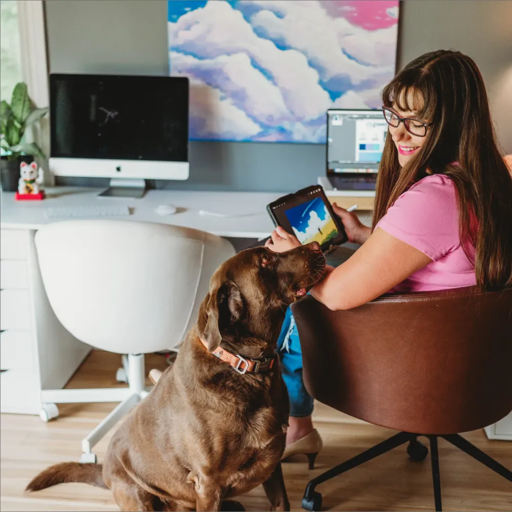 Leanne Jones drawing on a tablet at her home office desk while a chocolate Labrador looks up at her