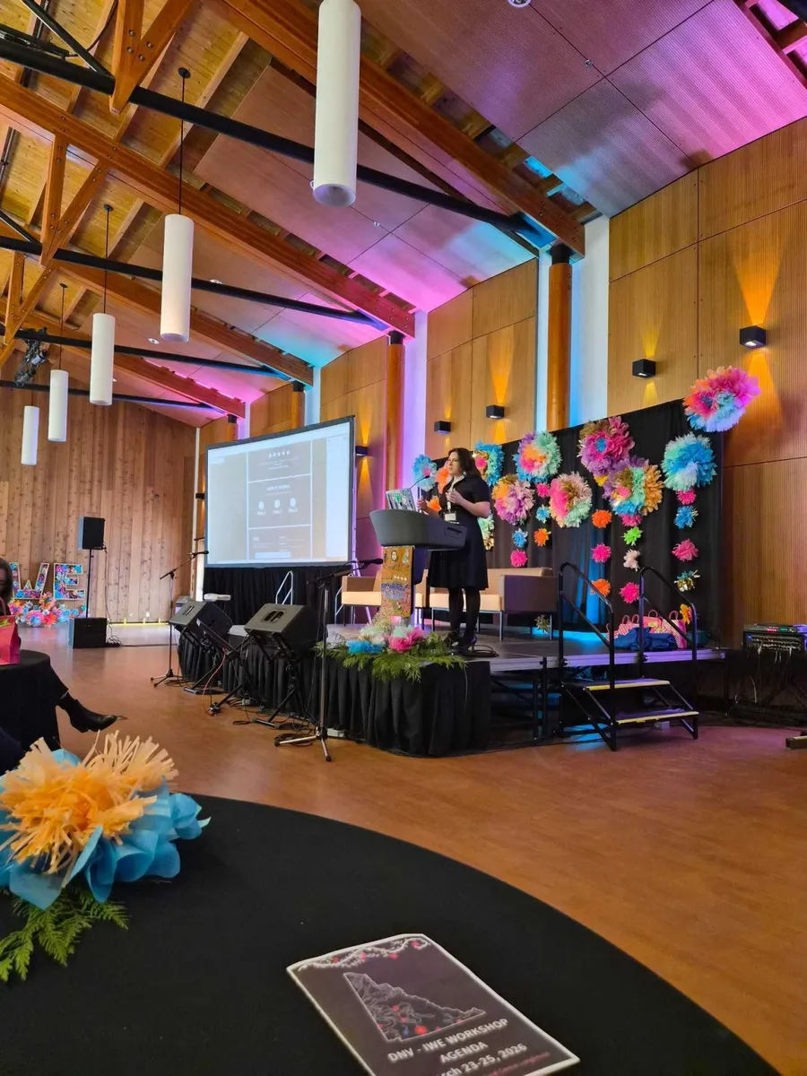 Leanne Jones presenting on stage beside a screen showing a website layout, with the event agenda on a table in the foreground