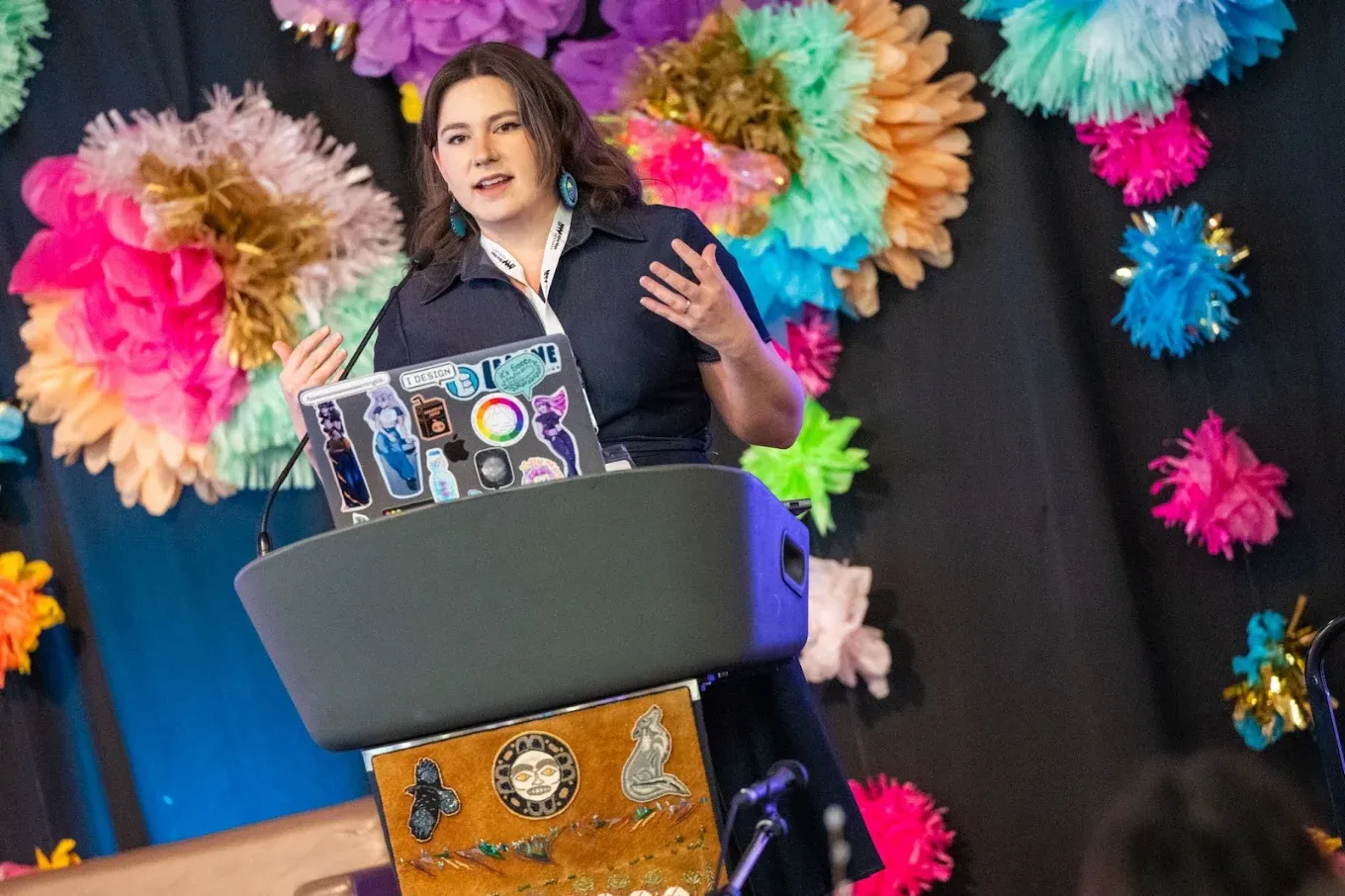 Leanne Jones speaking at a podium decorated with Indigenous artwork, in front of a wall of colourful paper flowers